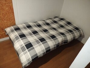 Uma cama de solteiro com colcha xadrez preta e branca em quarto com assoalho de madeira e paredes brancas.