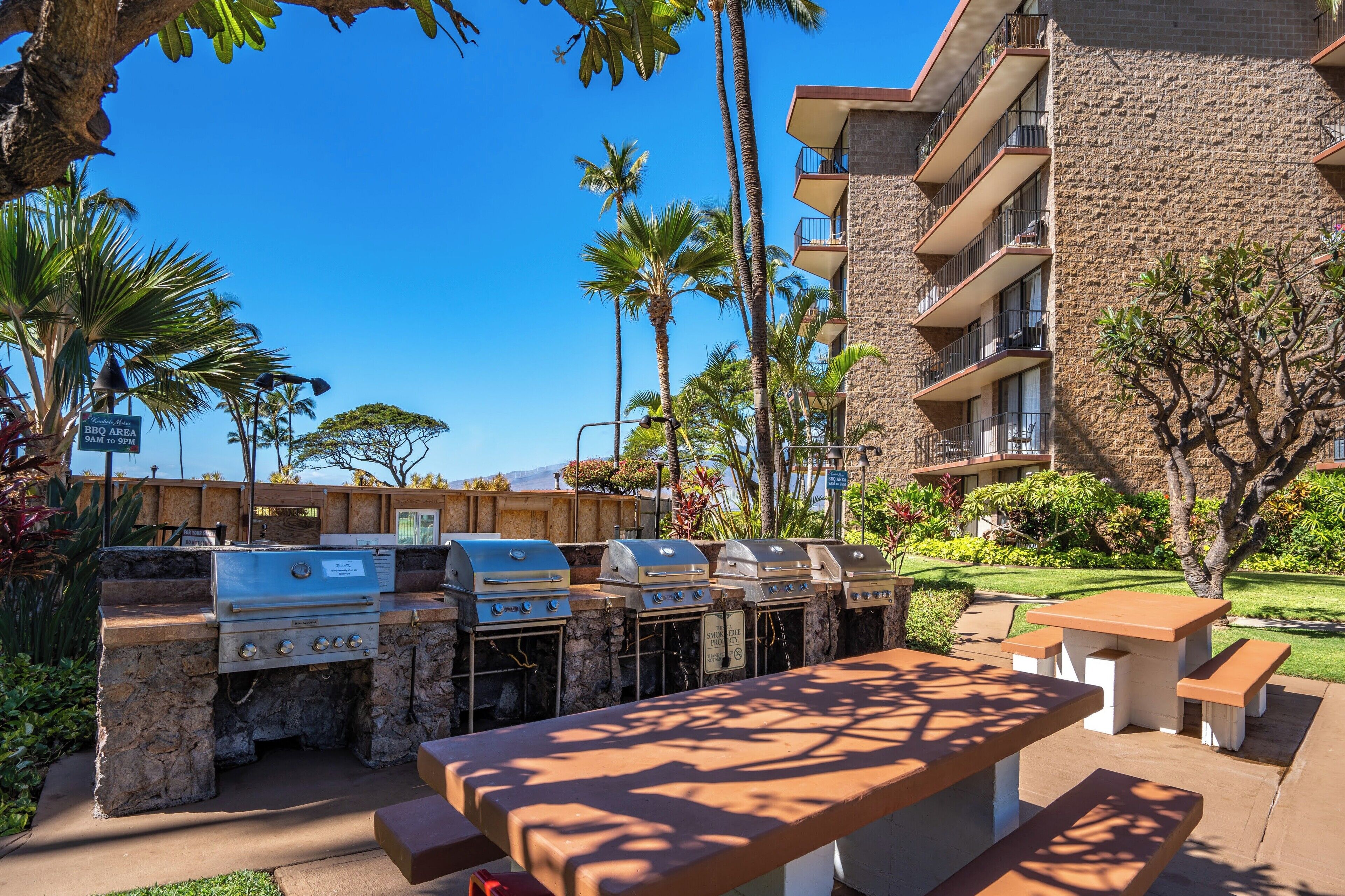 An outdoor barbecue area with stone-built grills, a wooden fence, and a view of a building with balconies.