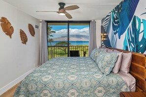 A bedroom with a large bed, a ceiling fan, and a view of a beach through a sliding door. - Hip Hawaiian Seaside Vistas (Kihei)