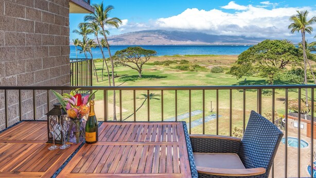A balcony with a wooden table, wicker chair, and a view of a golf course, palm trees, and a beach. - Hip Hawaiian Seaside Vistas (Kihei)