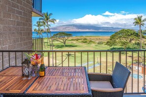Un balcón con mesa de madera, silla de mimbre y vista a un campo de golf, palmeras y una playa.