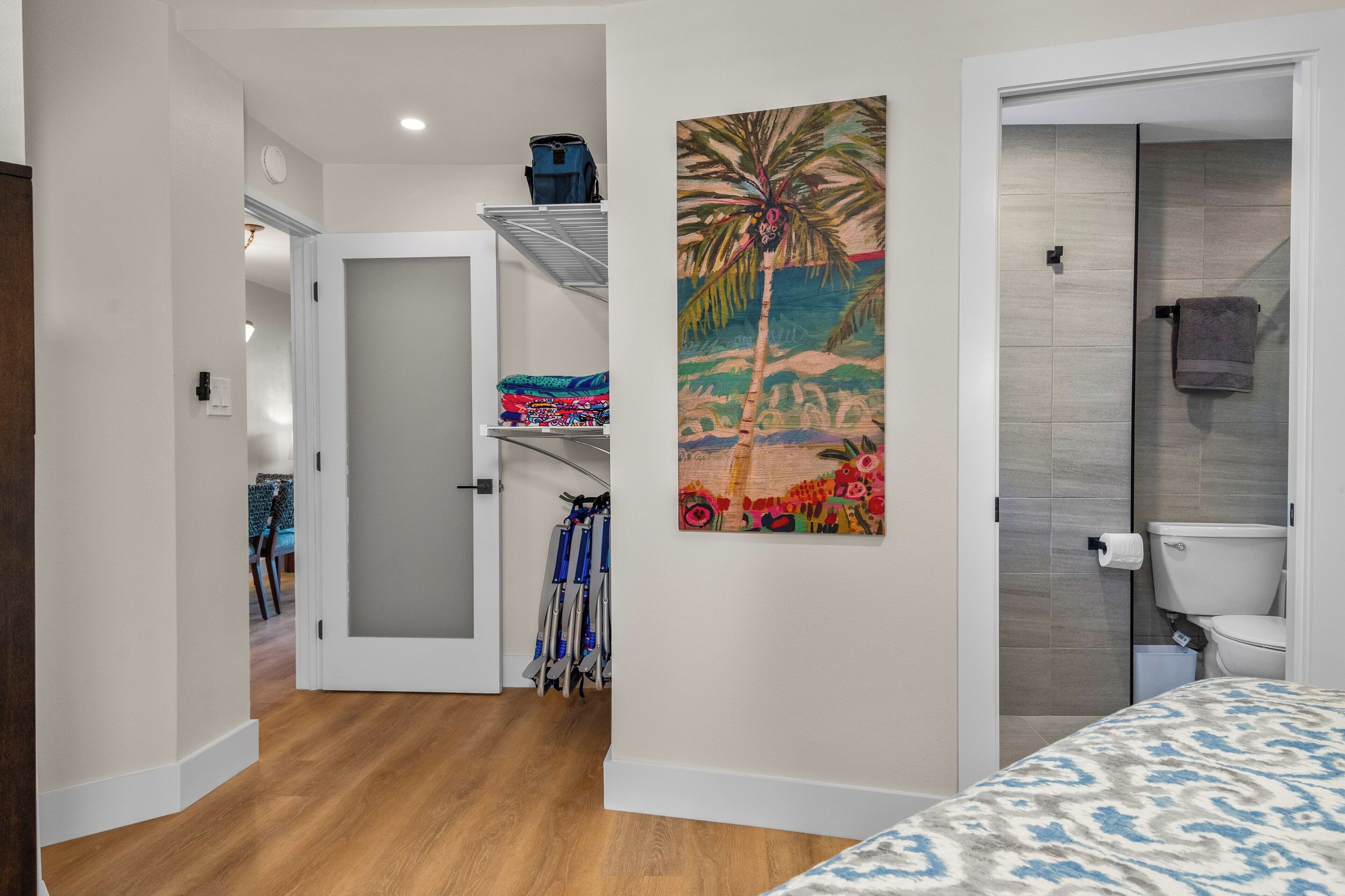 A hotel room with a wooden floor, a bed with a patterned bedspread, a painting of a tropical scene, a bathroom with a toilet and shower, and a hallway leading to another room.