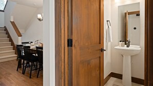 Eine Holztür führt zu einem Badezimmer mit Waschbecken und Spiegel, einem Essbereich mit Tisch und Stühlen sowie einer Treppe mit Holzstufen.