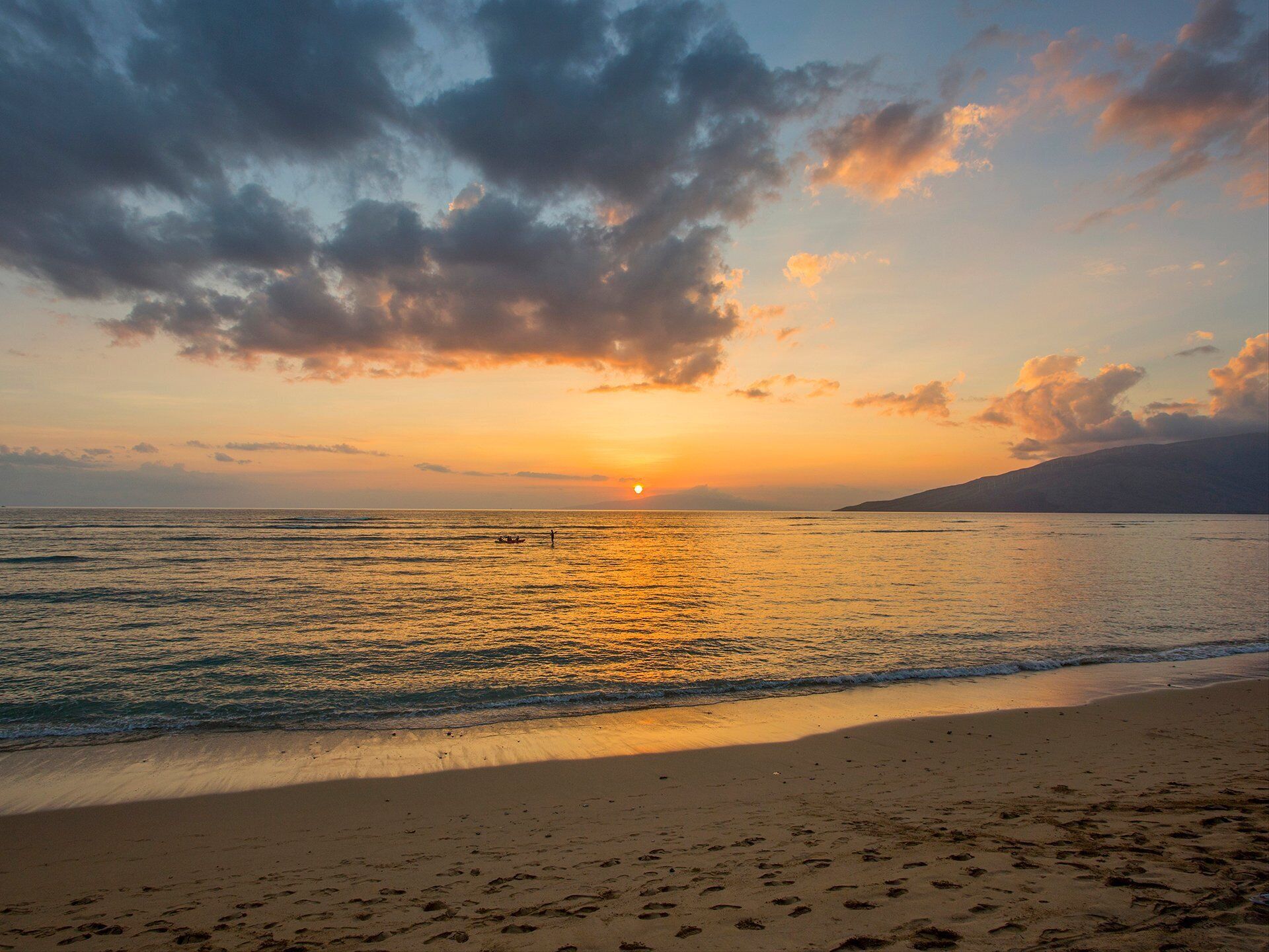 Una spiaggia con impronte che conducono all'acqua, un tramonto sullo sfondo e una montagna in lontananza.