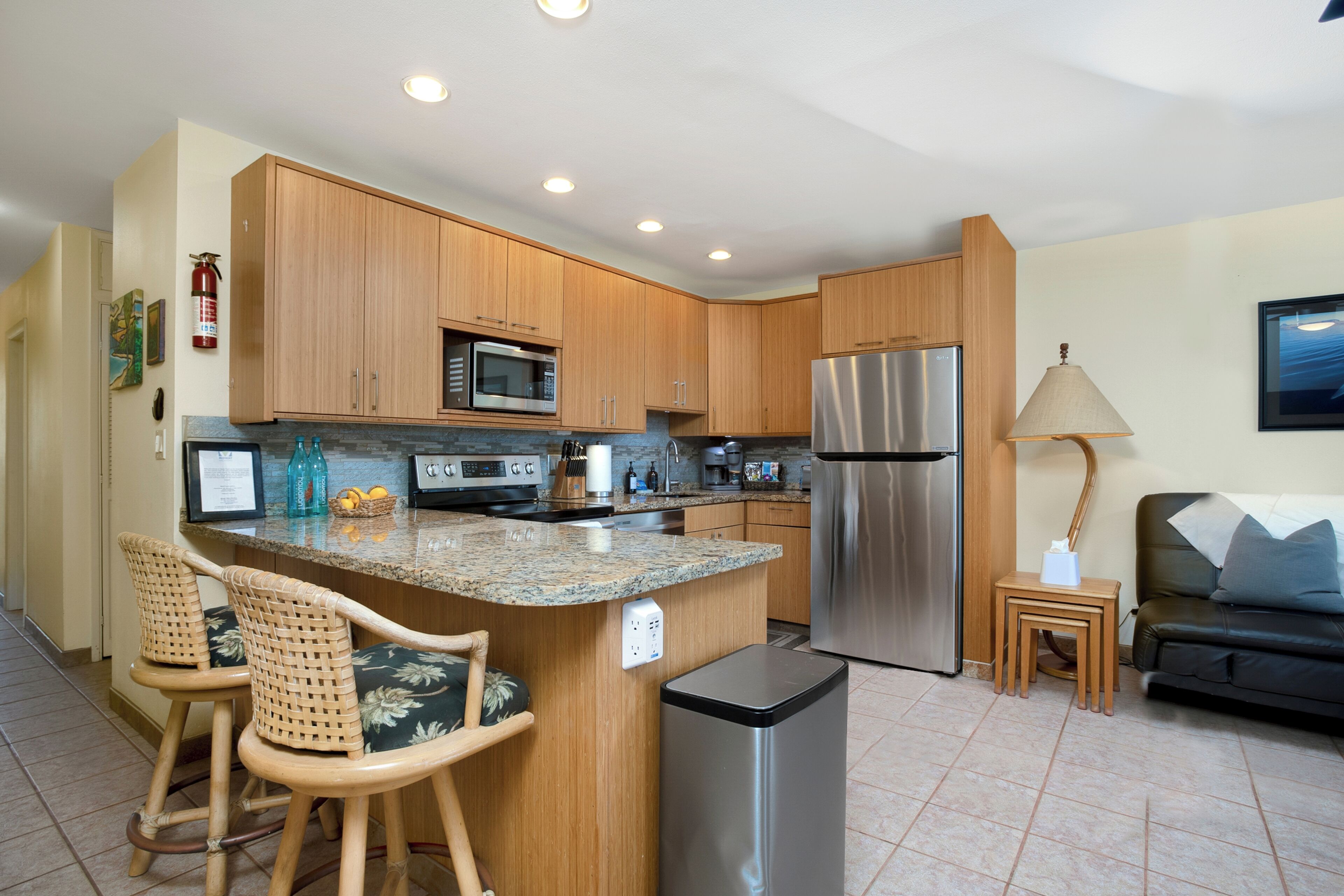 A modern kitchen with wooden cabinets, stainless steel appliances, a granite countertop, and a small dining area with wicker chairs.