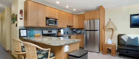 A modern kitchen with wooden cabinets, stainless steel appliances, a granite countertop, and a small dining area with wicker chairs.