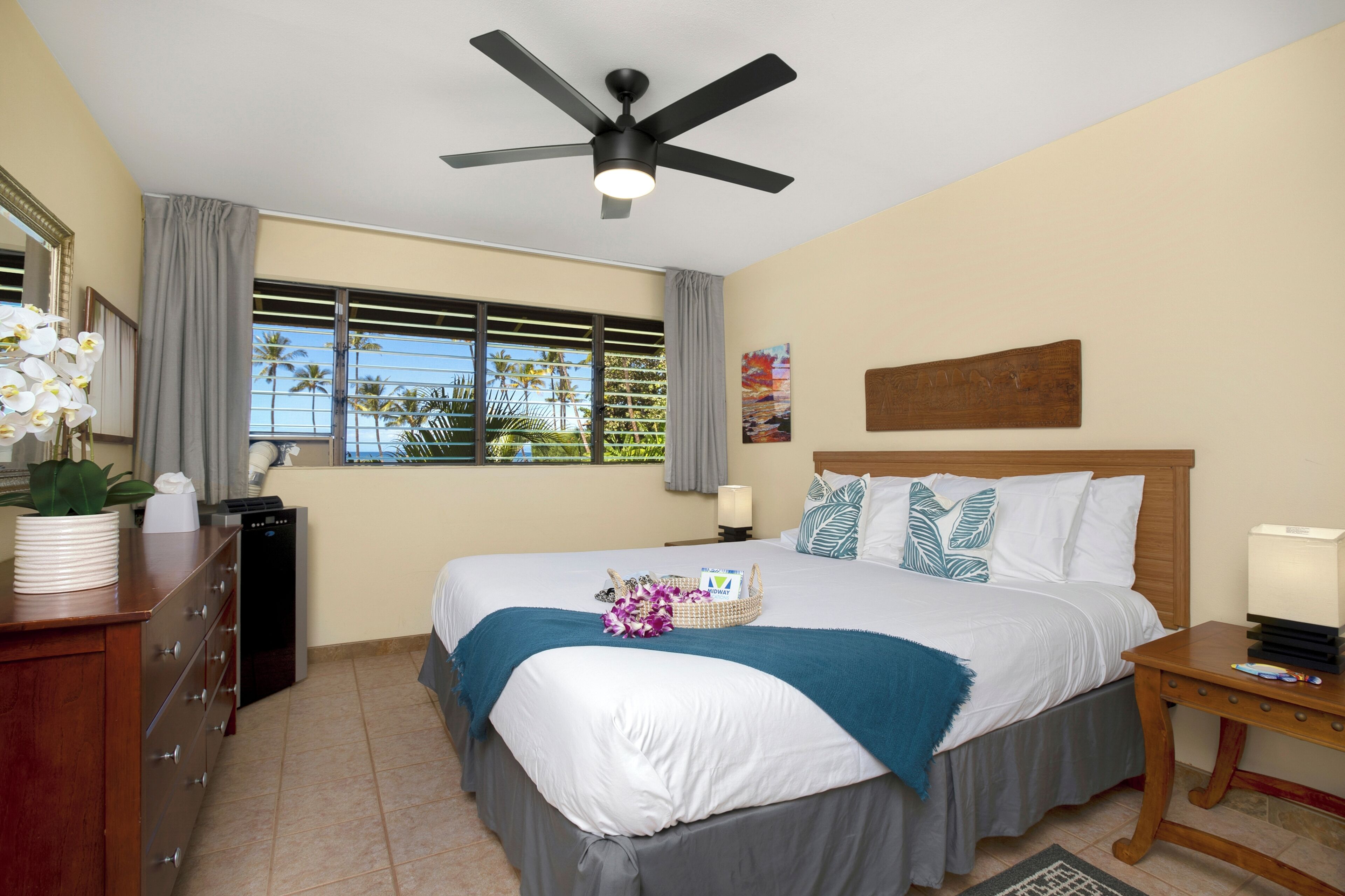 Una habitación de hotel con cama, ventilador de techo, ventana con cortinas, cómoda y mesita de noche.