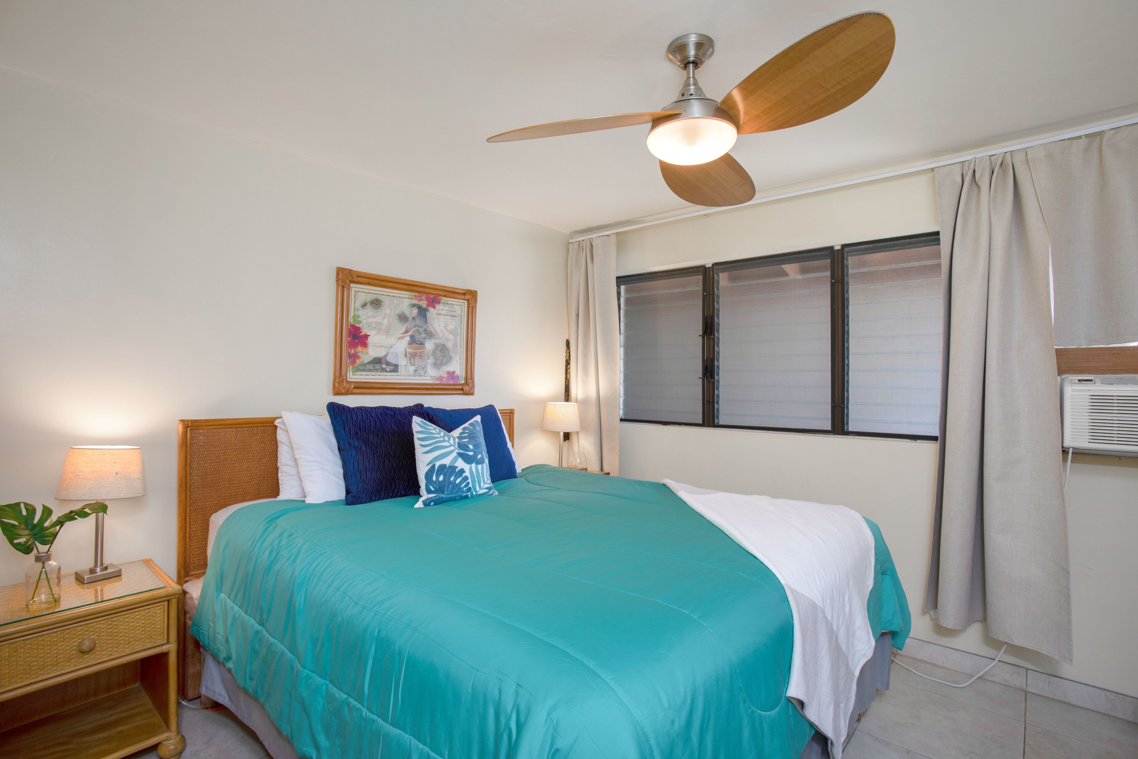 A bedroom with a bed, a ceiling fan, a nightstand with a lamp, a window with curtains, and a wall-mounted air conditioner.