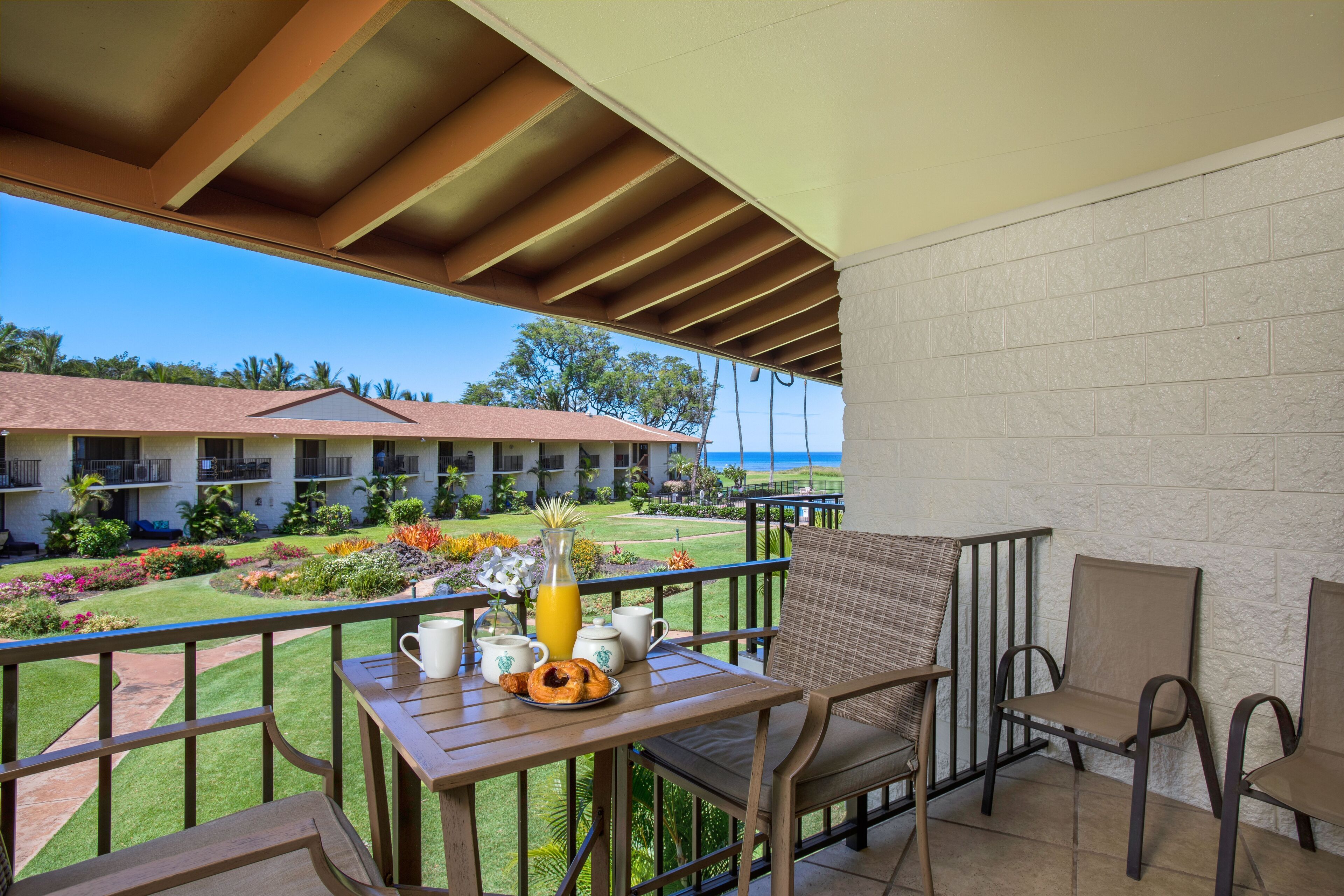 A balcony with a table set for breakfast, overlooking a hotel courtyard with a pool and a view of the ocean.