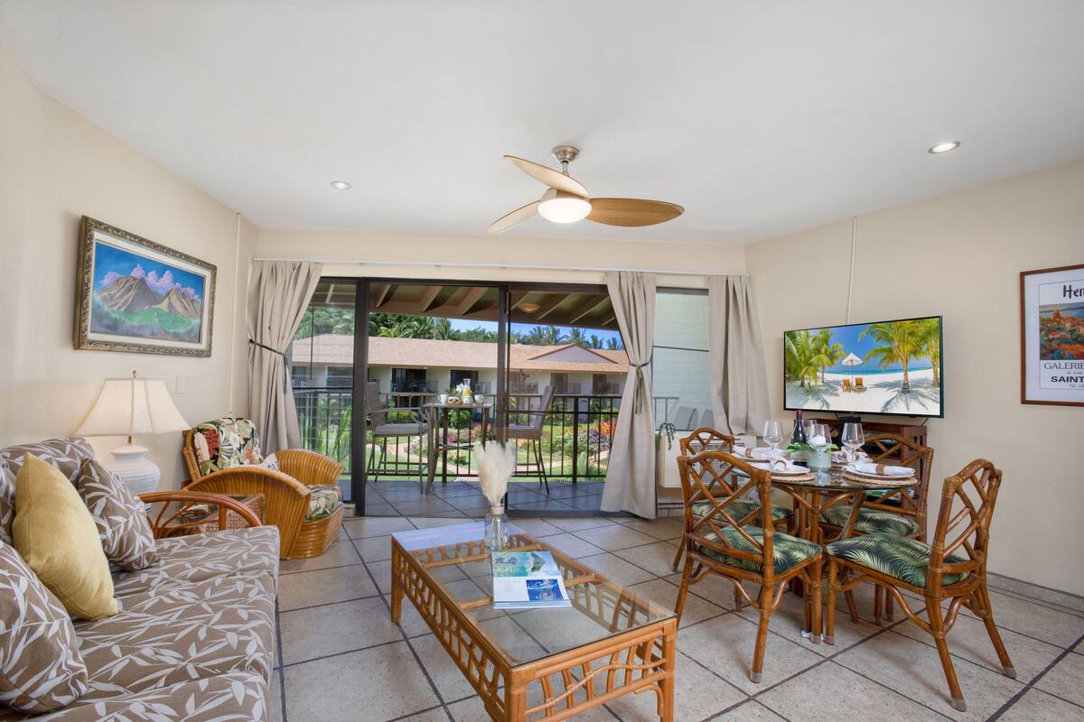A spacious living area with a sofa, dining table, and chairs, a ceiling fan, and a TV.