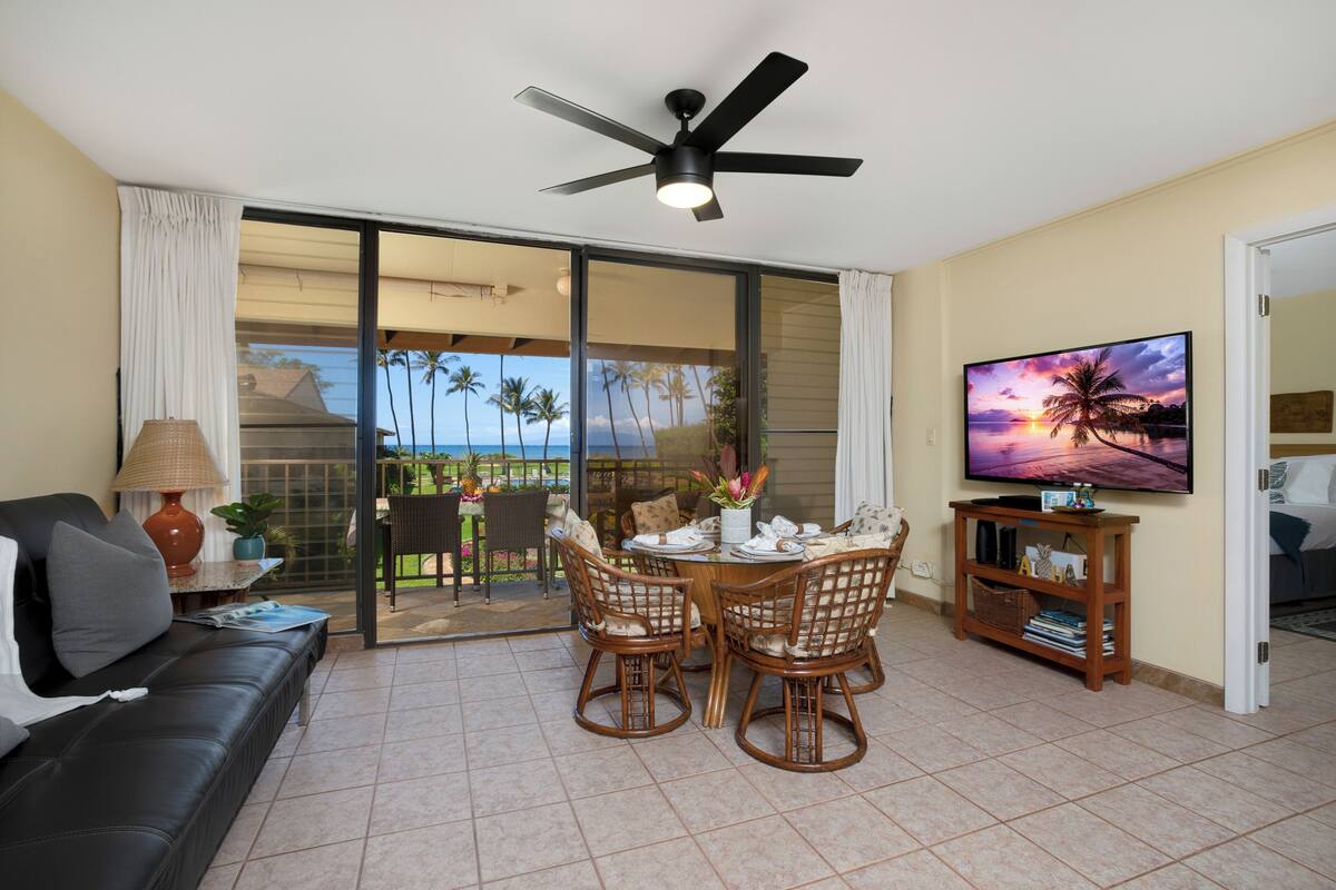 A living room with a black leather sofa, a dining table with wicker chairs, a ceiling fan, a flat-screen TV, and a sliding glass door leading to a balcony with a view of palm trees and the ocean.