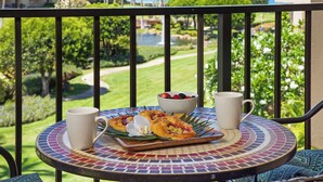 A round table with a mosaic tablecloth, two white mugs, a bowl of fruit, and a plate with pastries on a balcony overlooking a garden and a body of water. - Kamaole Sands Condos Close to Beach with Parking (Kihei)