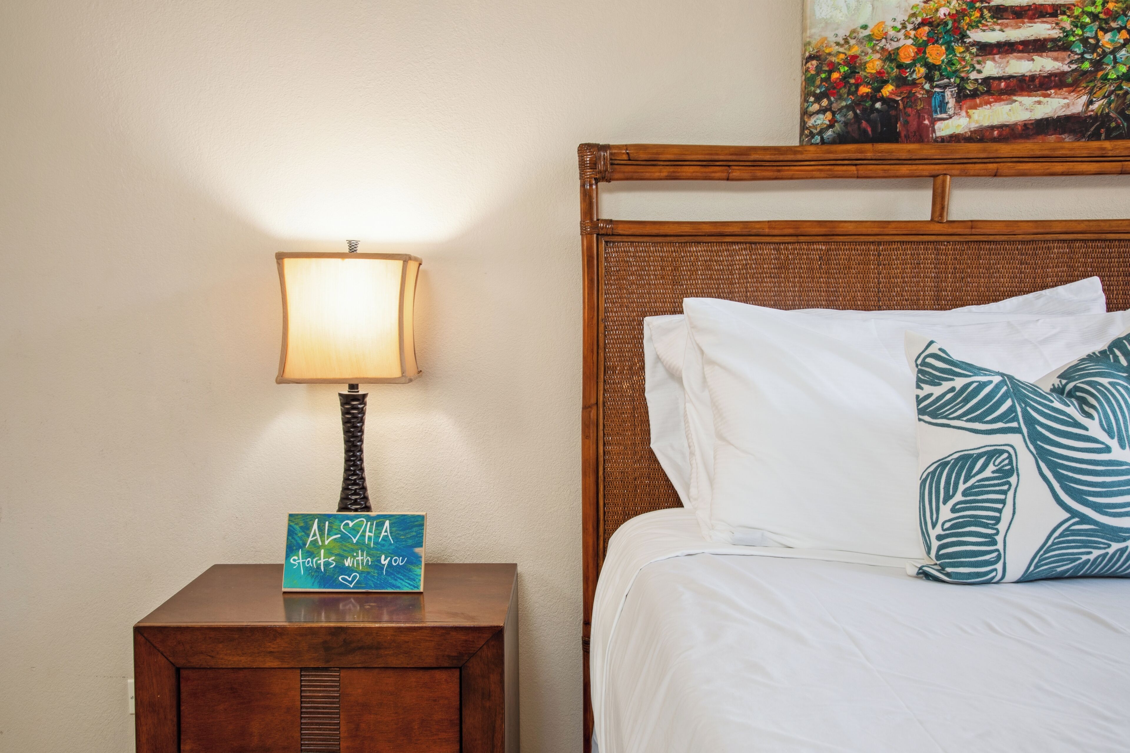 Una cama bien hecha con sábanas blancas y almohadas estampadas con hojas, una lámpara de noche y una mesita de noche de madera con un cartel que dice "ALWAYS starts with you".