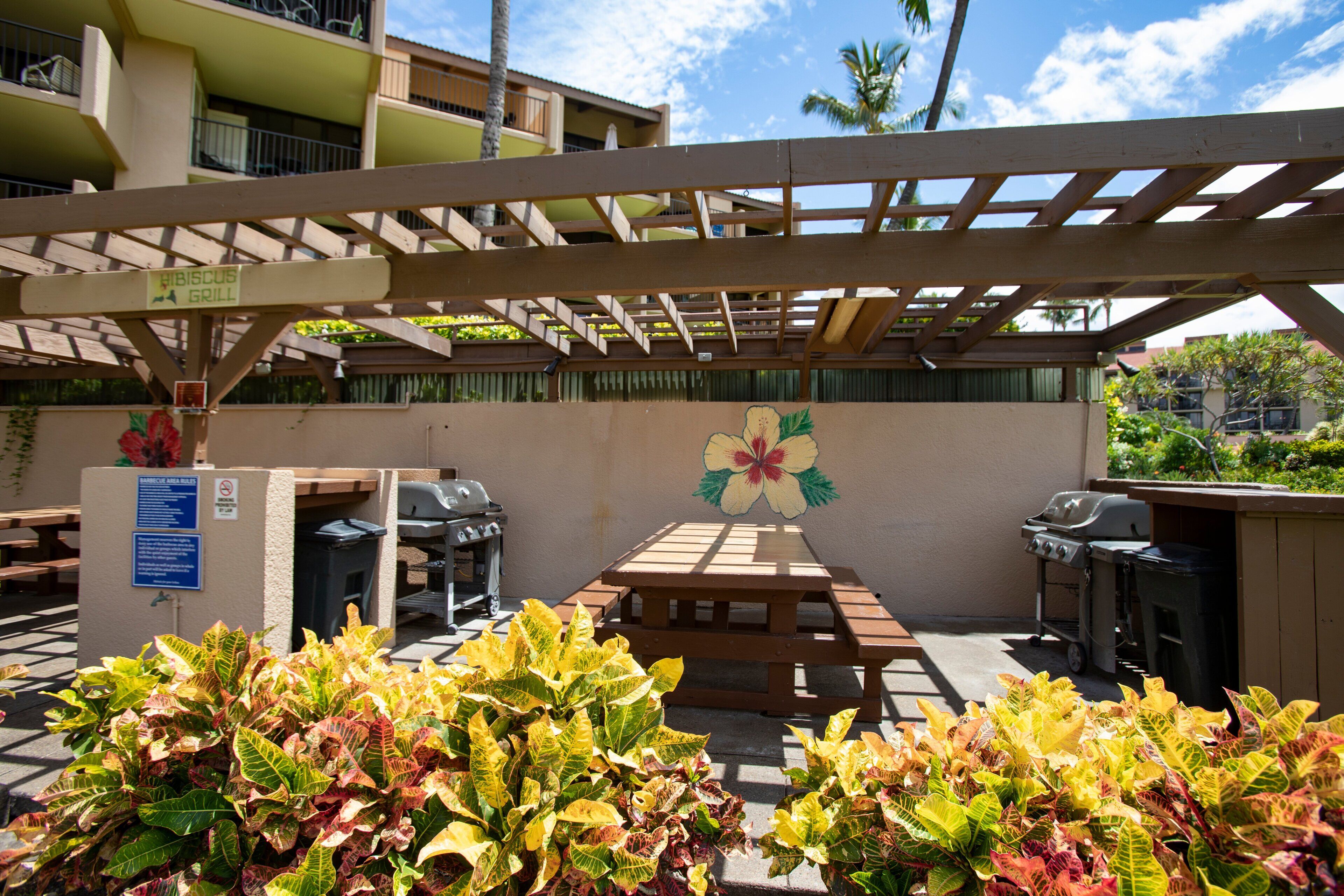 An outdoor patio area with a wooden pergola, a hibiscus flower mural, a grill, and a trash bin.