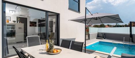 A modern outdoor dining area with a pool, a table with chairs, and a large umbrella.