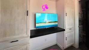 Un téléviseur posé sur une étagère noire, dans une pièce où les meubles sont blancs et où se trouve un tapis rose.