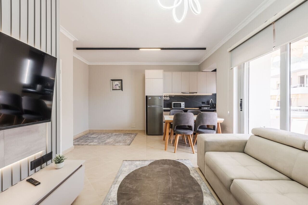 A modern living room with a flat-screen TV, a sofa, a dining area with chairs, and a kitchen in the background.