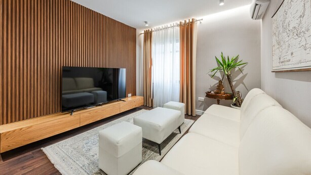 A modern living room with a flat-screen TV, white sofas, a wooden accent wall, and a large window with curtains.