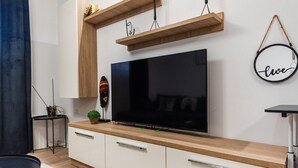 Ein modernes Wohnzimmer mit einem großen Flachbildfernseher an einer Holzkommode, einem kleinen Beistelltisch mit Pflanze und einem Wandregal mit Dekoration.