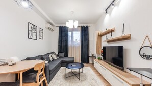 Ein modernes Wohnzimmer mit einer Eckcouch, einem Esstisch mit Stühlen, einem Flachbildfernseher und einem großen Fenster mit Vorhängen.