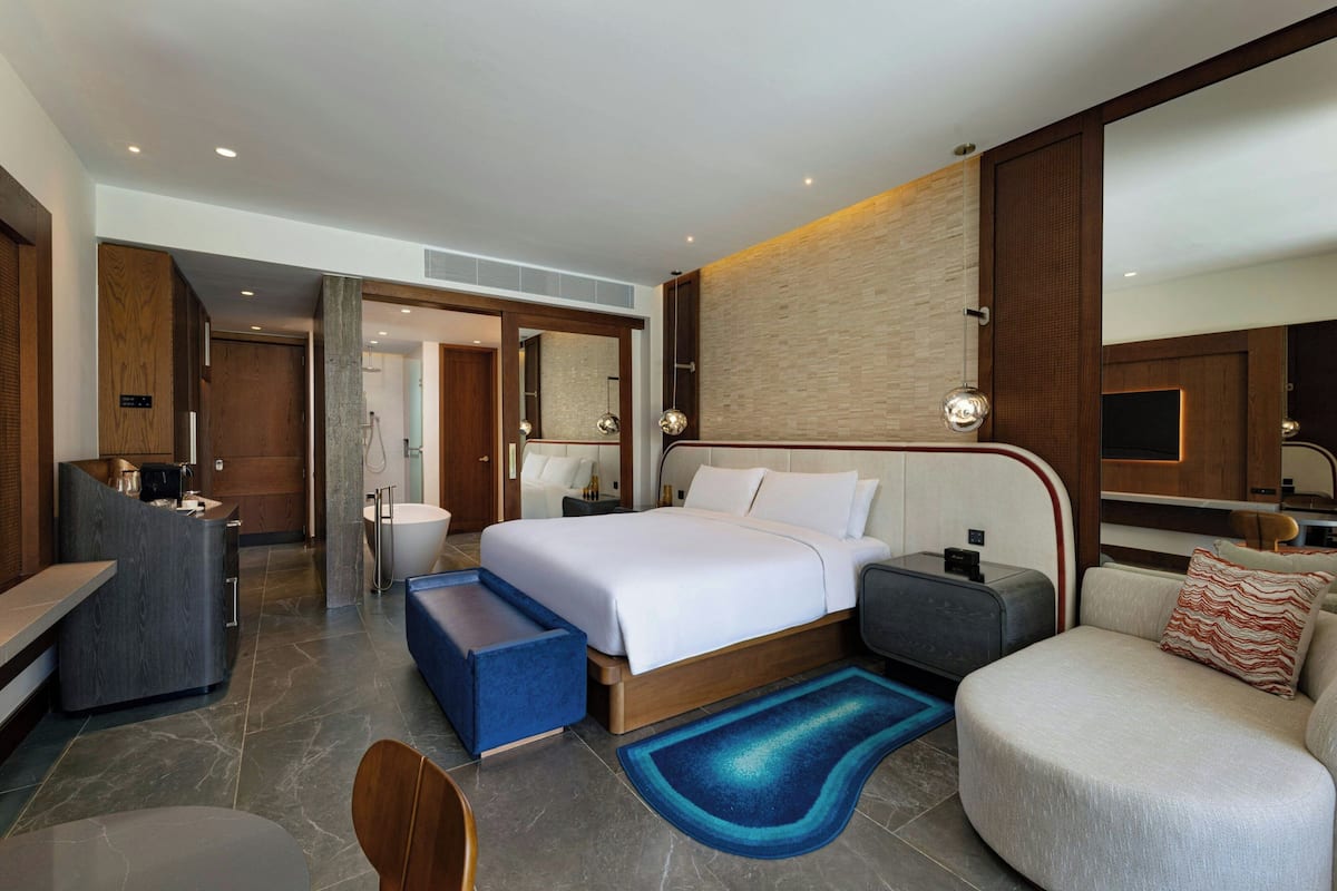 A modern hotel room with a large bed, a seating area, a desk, and a bathroom.