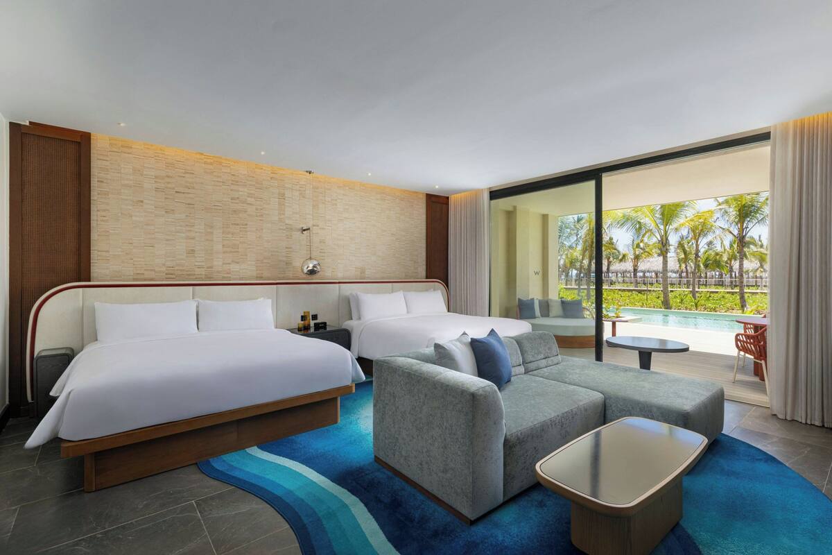 A hotel room with two beds, a sofa, a coffee table, and a view of a pool and palm trees.