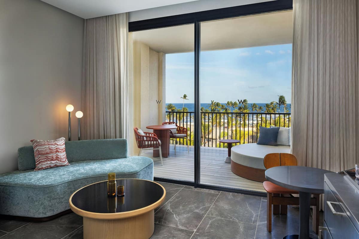 A modern hotel room with a curved sofa, a round coffee table, a dining table, and a balcony with a view of the ocean.