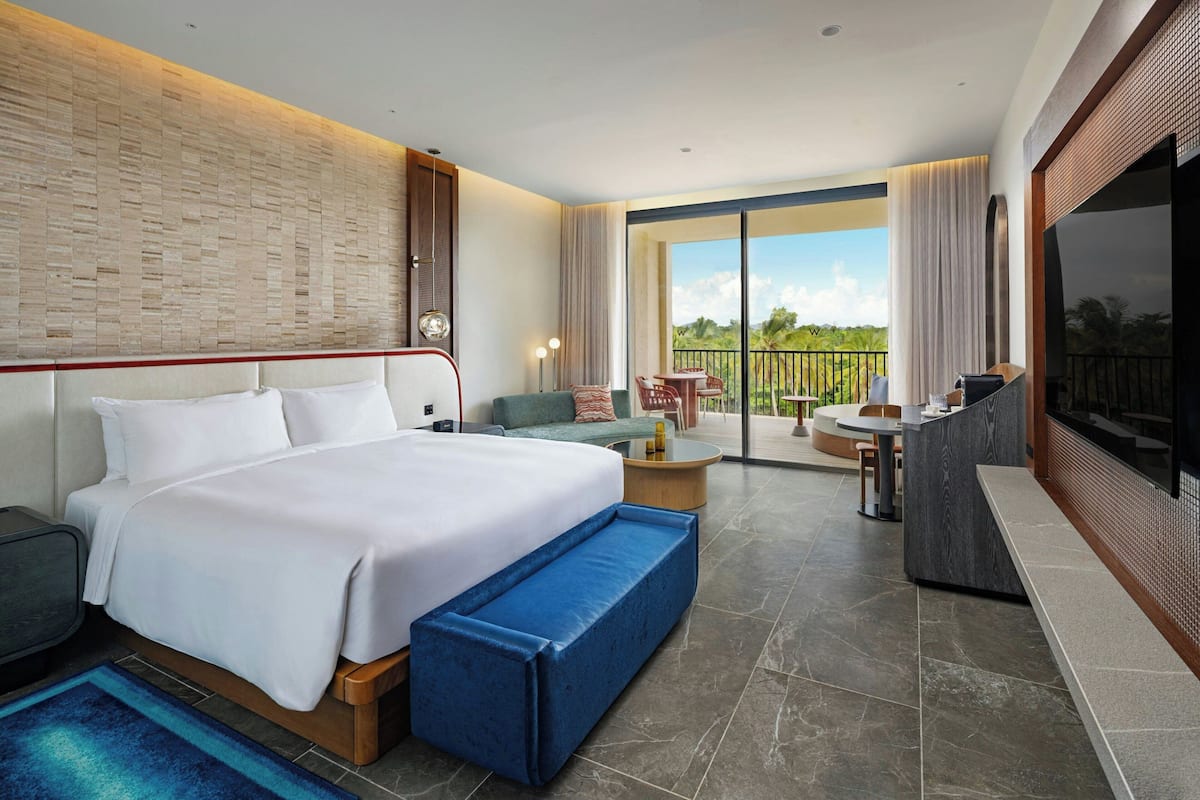 A modern hotel room with a large bed, a seating area with a table, a TV, and a balcony with a view.