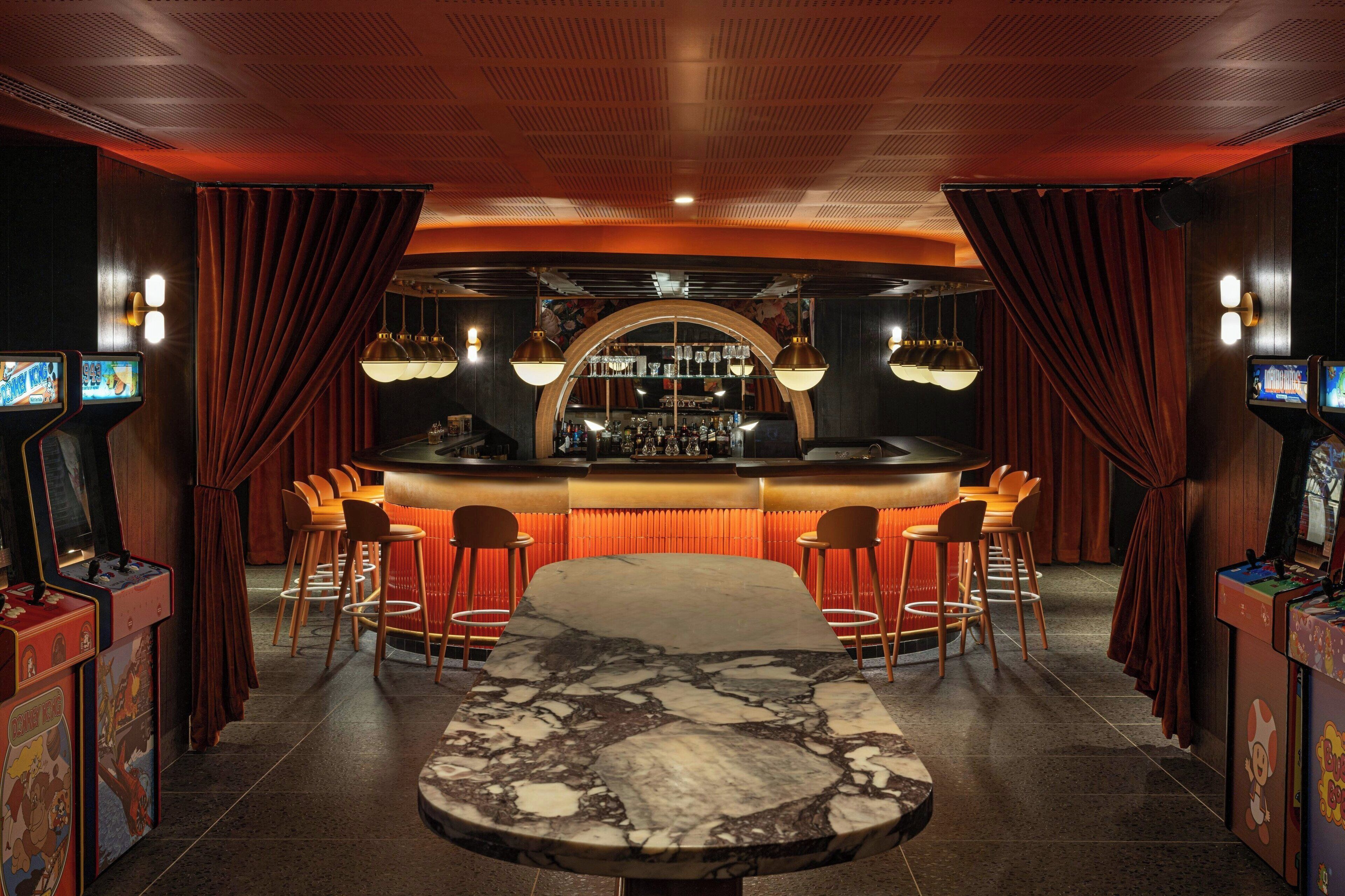 Uma área de bar com mesa de mármore, banquetas e máquinas de jogos.