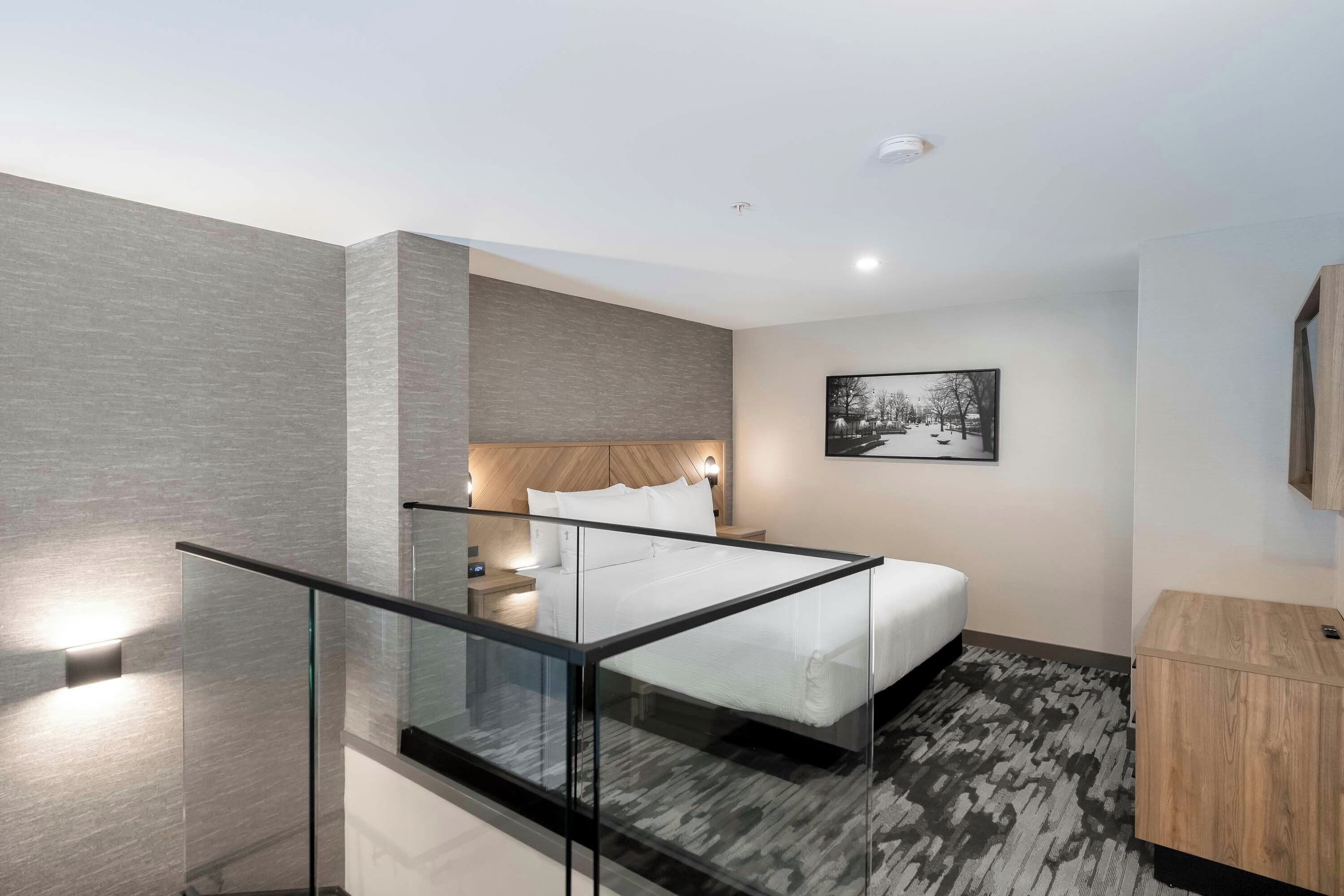 Une chambre d’hôtel moderne avec un balcon en verre, un lit recouvert de draps blancs, une tête de lit en bois et un tableau encadré au mur.