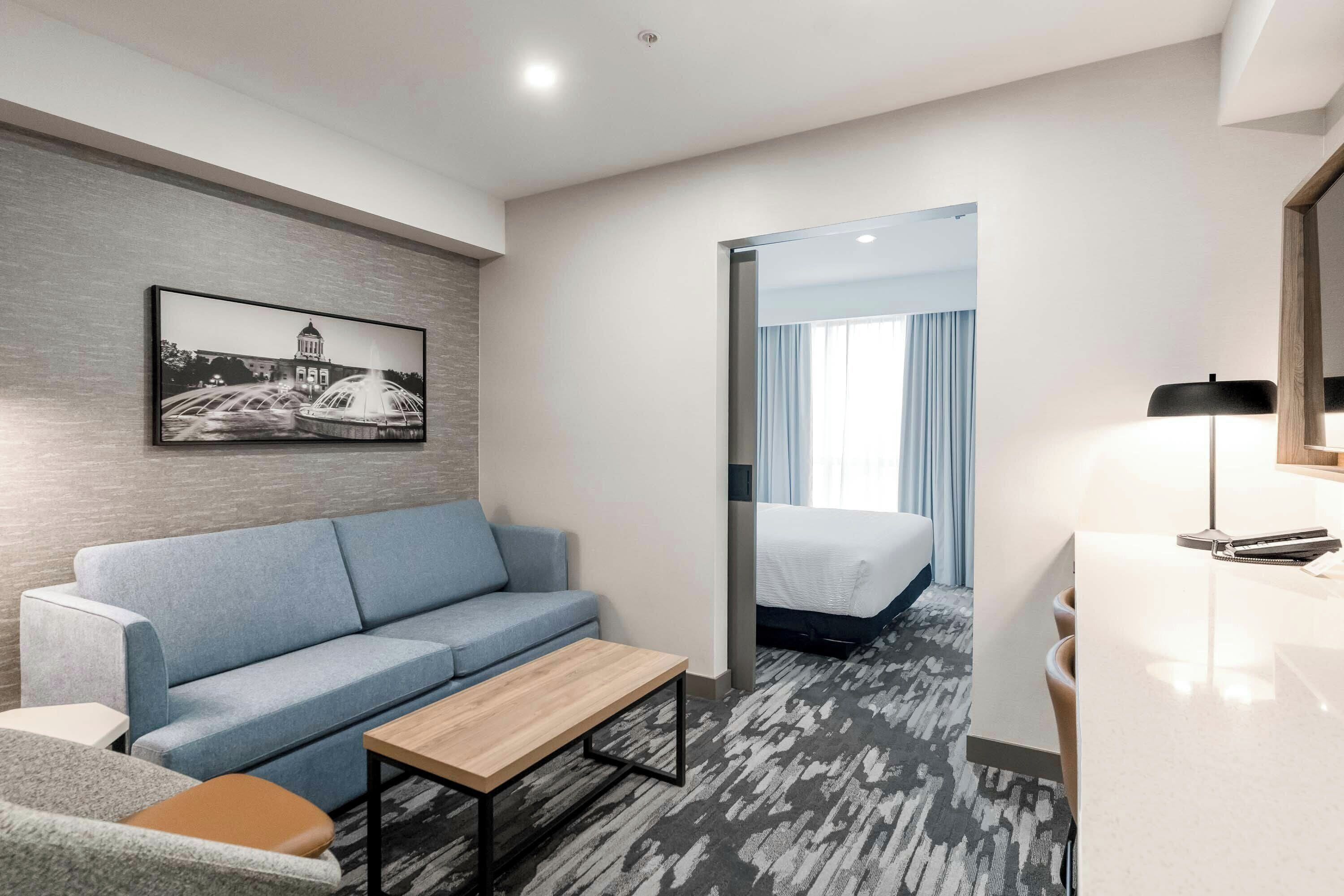 A hotel room with a sofa, a small table, a bed, a desk with a lamp, and a picture on the wall.