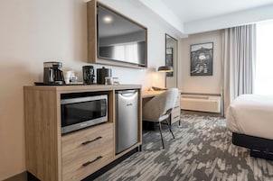 Habitación de hotel con cama, escritorio con silla, minibar, cafetera, microondas y un espejo grande.