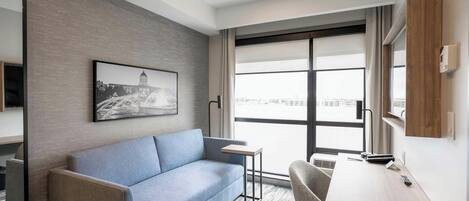 A modern hotel room with a sofa, a small table, a desk, a chair, a window with a view, and a mounted picture on the wall.