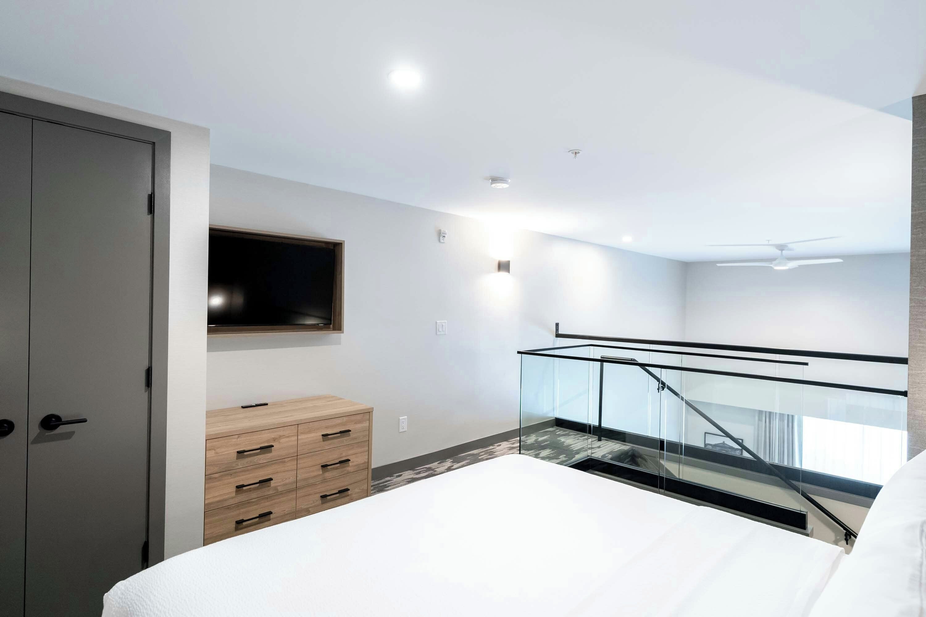 Un dormitorio moderno con cama, televisor montado en la pared, cómoda de madera y baranda de vidrio con vistas a un balcón.