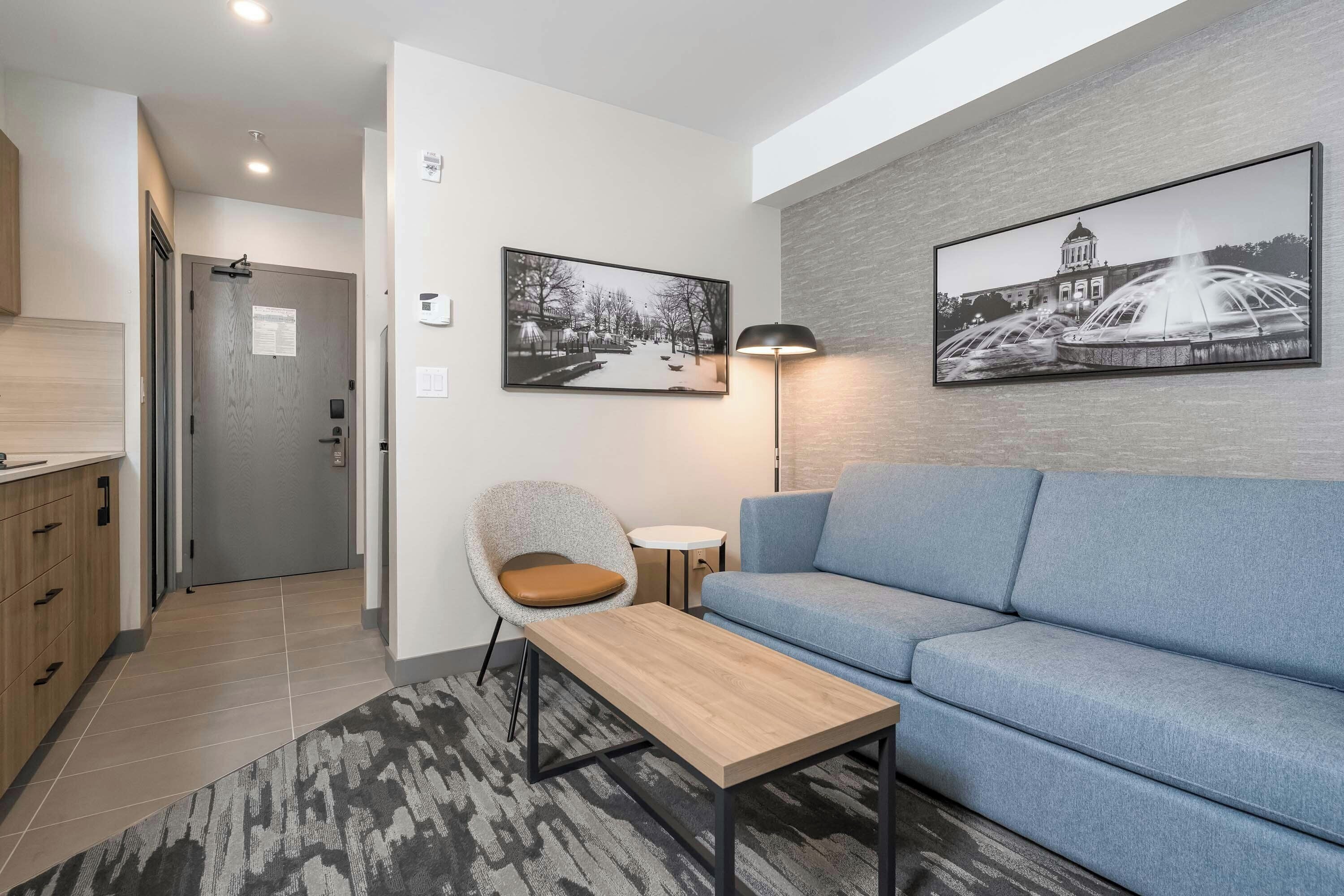 A modern living room with a sofa, a coffee table, and a chair. There are two framed pictures on the wall, a lamp, and a door with a sign.