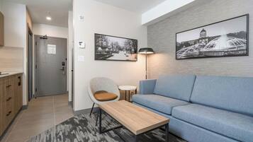 A modern living room with a sofa, a coffee table, and a chair. There are two framed pictures on the wall, a lamp, and a door with a sign.