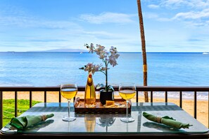 Une table avec deux verres de vin blanc, une bouteille de vin et un vase rempli de fleurs blanches, donnant sur une plage et l’océan.