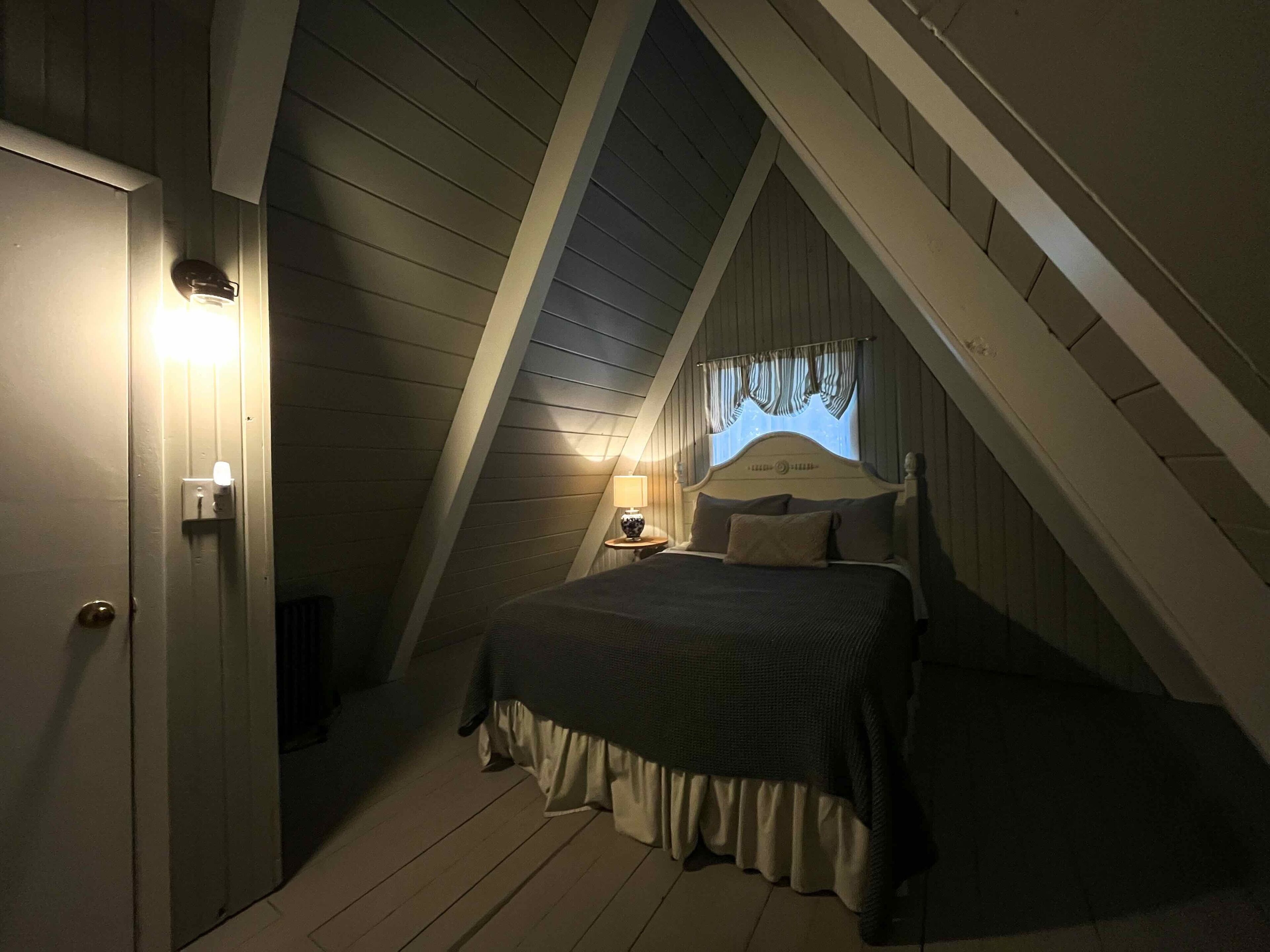 Ein Schlafzimmer mit schrägem Dach, einem Bett mit Kopfteil, einem Nachttisch mit Lampe und einer Tür mit Lichtschalter.