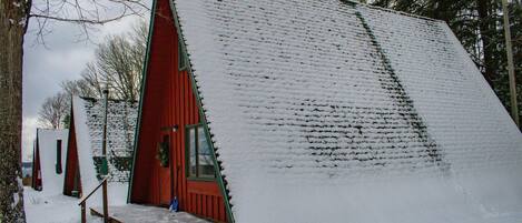 Une cabane rouge, dotée d’un toit à forte pente recouvert de neige, entourée d’arbres.