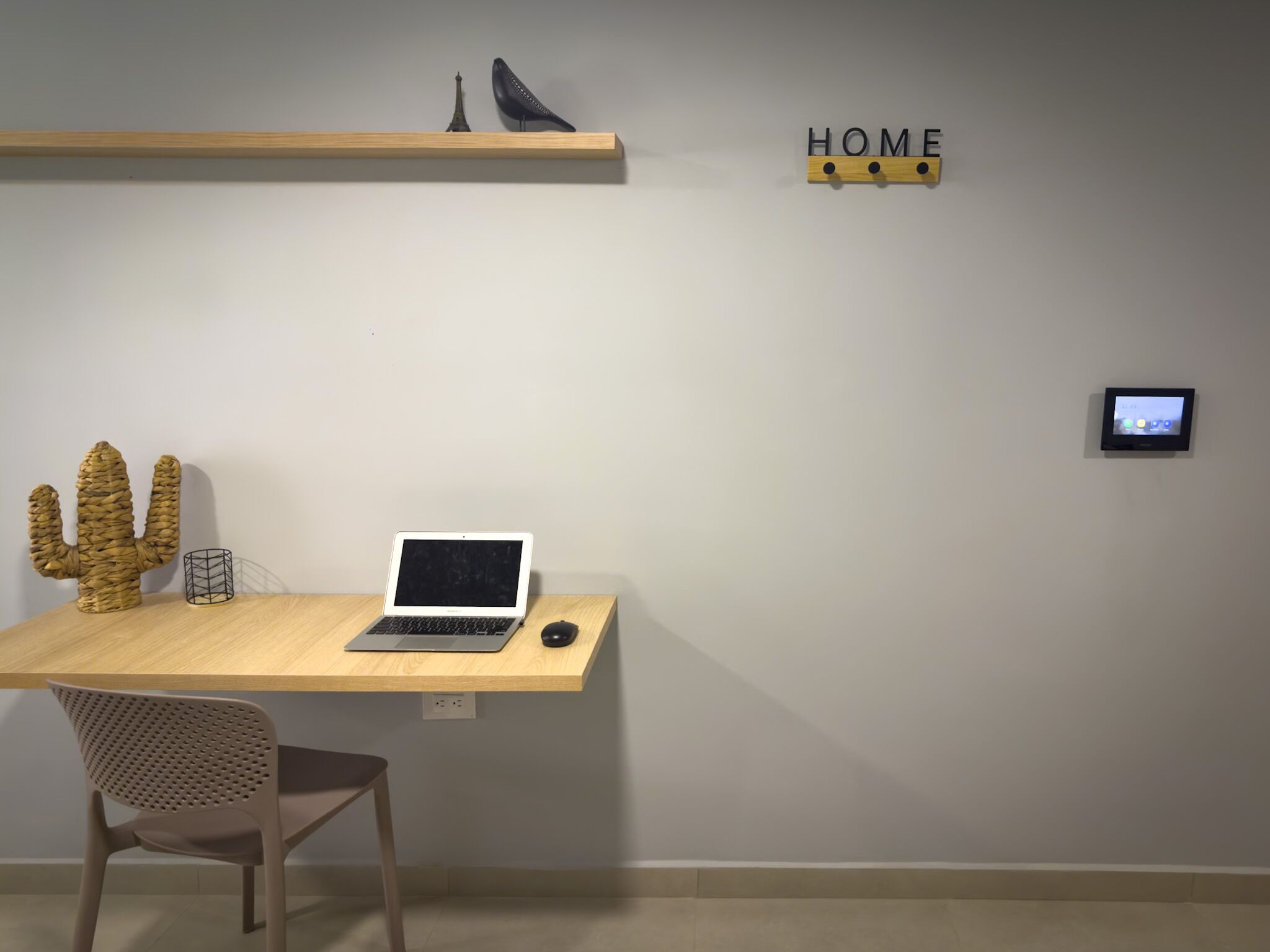 A minimalist workspace with a laptop on a wooden desk, a cactus sculpture, a chair, and a wall-mounted thermostat.