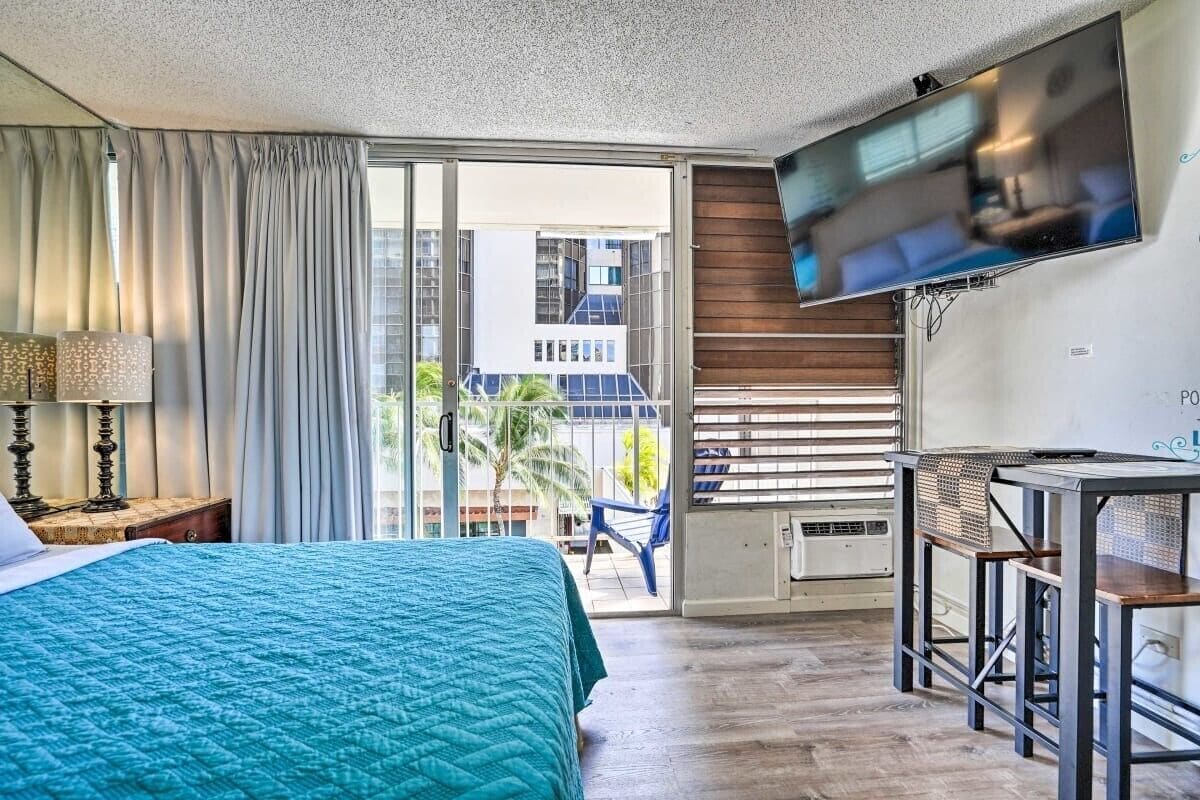 A hotel room with a bed, a TV mounted on the wall, a balcony with a view of buildings, and a small table with chairs.