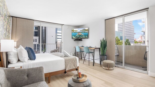 A modern hotel room with a bed, a desk, a TV, and a balcony with a city view. - Marine Surf by Tropical Escapes Realty (Honolulu)
