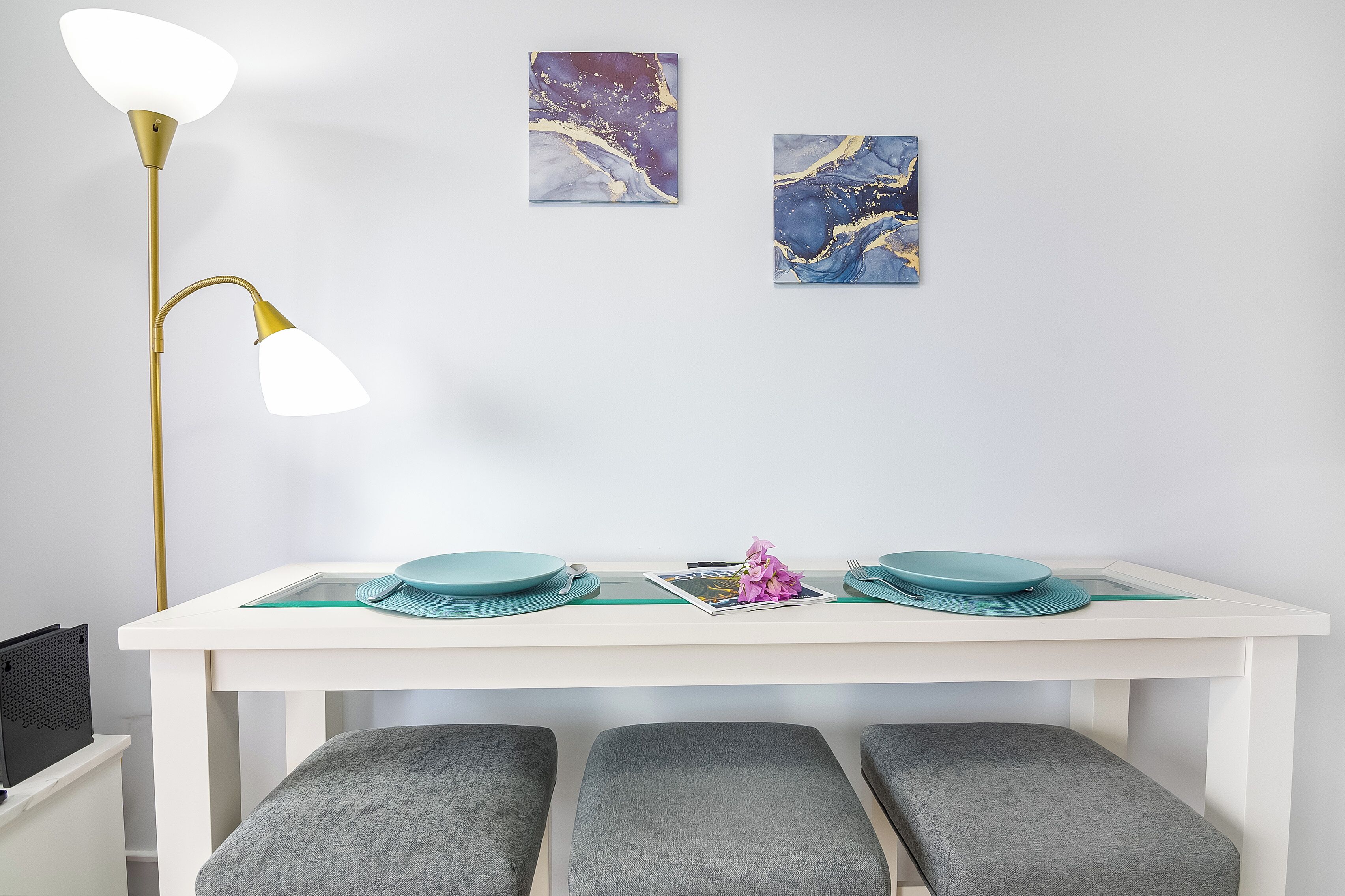 Un coin repas avec une table blanche, des bancs gris rembourrés, un lampadaire et deux tableaux abstraits accrochés au mur.
