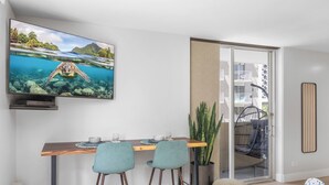 A modern living room with a wooden dining table, two blue chairs, a TV displaying a sea turtle, and a sliding glass door leading to a balcony with a hanging chair. - Marine Surf by Tropical Escapes Realty (Honolulu)