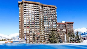 Ein mehrstöckiges Hotel mit zahlreichen Balkonen, im Hintergrund ein Skigebiet und ein strahlend blauer Himmel.