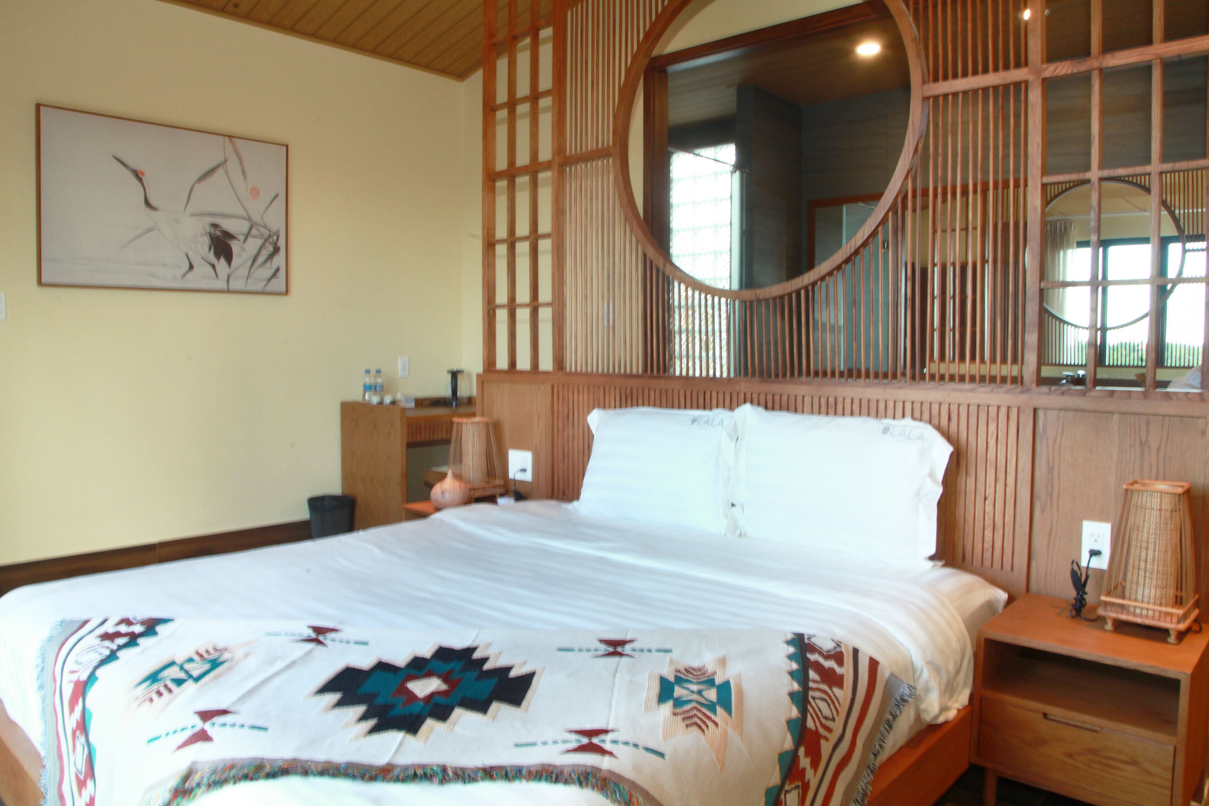 A bedroom with a large bed, a wooden headboard with a mirror, a bedside table with a lamp, and a wall-mounted artwork.