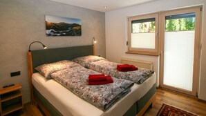 A double bed with floral bedding, a nightstand, a wall-mounted lamp, a painting of a landscape, and a window with blinds.