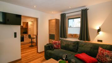 Ein modernes Wohnzimmer mit einem grünen Sofa, einem Holztisch und einer durchgehenden Tür erkennbaren Küche.
