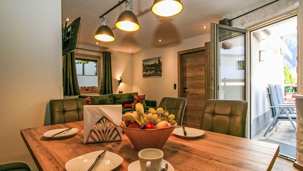 A dining area with a wooden table, chairs, a fruit bowl, plates, cutlery, and a cup.