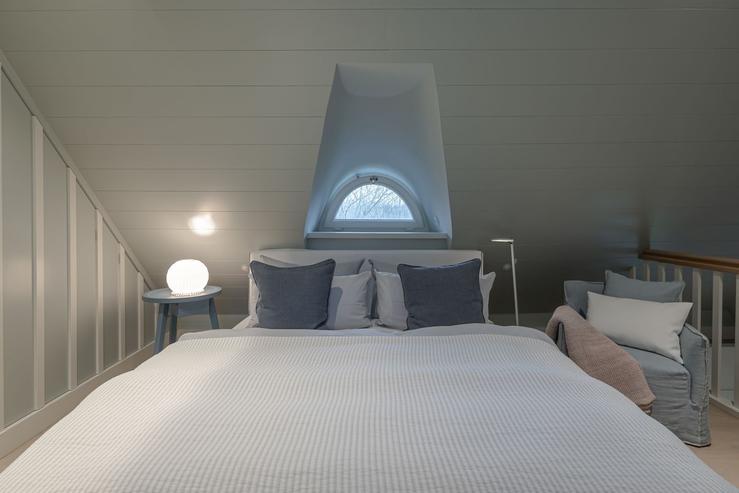Une chambre avec un grand lit, une lucarne, une table de chevet avec une lampe et une chaise.